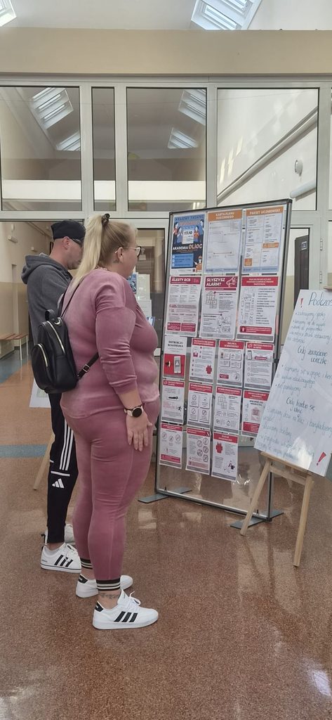 Na zdjęciu przedstawiono uczestników Krajowego Treningu Rodzinnego – Akademii Ochrony Ludności i Obrony Cywilnej (OLiOC), którzy zapoznają się z wystawą edukacyjną przygotowaną na szkolnym korytarzu.
Na specjalnej tablicy informacyjnej zaprezentowano materiały dotyczące zasad bezpieczeństwa, reagowania na sygnały alarmowe i Alerty RCB, przygotowania rodzinnego planu bezpieczeństwa, wyboru miejsca schronienia oraz właściwego postępowania w sytuacjach kryzysowych. Obok wystawy znajduje się również plakat wykonany przez uczniów, przedstawiający zasady zachowania podczas alarmu.
