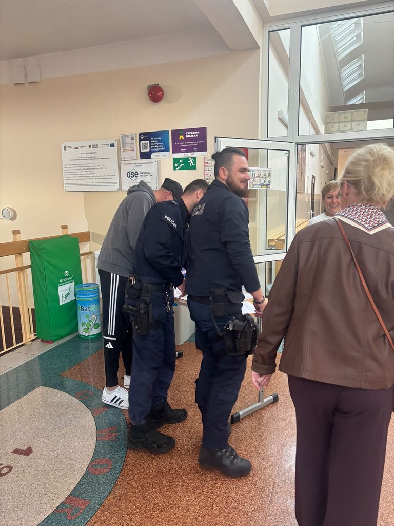 Na zdjęciu przedstawiono spotkanie z funkcjonariuszami Policji w ramach Krajowego Treningu Rodzinnego – Akademii Ochrony Ludności i Obrony Cywilnej (OLiOC).
Na szkolnym korytarzu widoczni są funkcjonariusze Policji oraz uczestnicy wydarzenia, którzy zgromadzili się przy stanowisku informacyjnym. Spotkanie stanowiło okazję do bezpośredniego kontaktu z przedstawicielami służb mundurowych oraz podkreślenia ich ważnej roli w zapewnianiu bezpieczeństwa mieszkańcom.