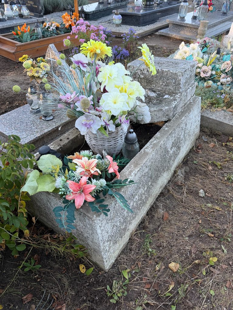 W centrum zdjęcia znajduje się betonowy nagrobek. Jest świeżo uprzątnięty. Widać dwa bukiety sztucznych, kolorowych kwiatów. Na nagrobku nie ma tablicy z imieniem i nazwiskiem. Dookoła rozciągają się inne nagrobki.
