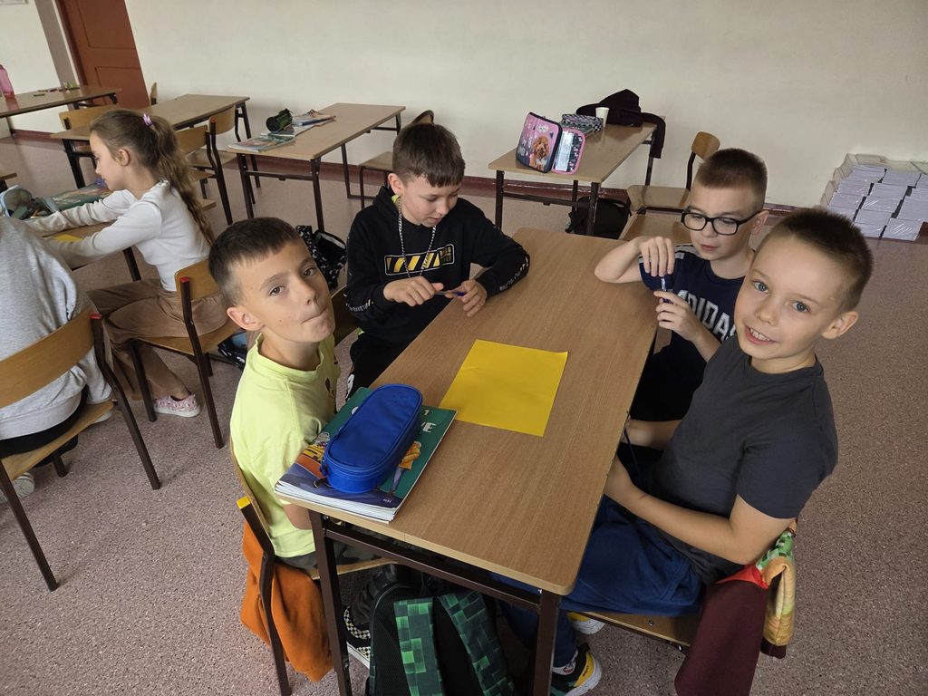 Na zdjęciu widoczna jest grupa uczniów pracujących przy szkolnej ławce. Czterech chłopców siedzi wokół stolika, na którym znajduje się żółta kartka, przybory szkolne, zeszyty i piórnik. Dwóch uczniów spogląda w stronę aparatu, a pozostali zajmują się przyborami. W tle widać również uczennice pracujące przy innych ławkach.
Fotografia przedstawia atmosferę szkolnej współpracy i zaangażowania w zajęcia.