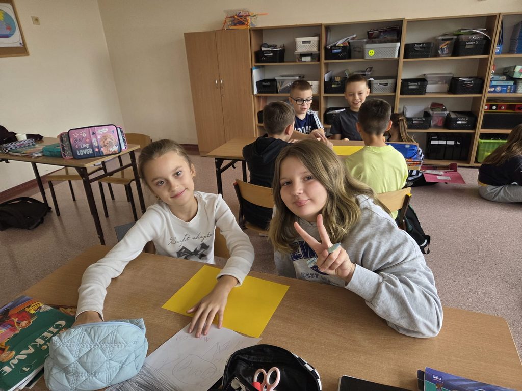 Na zdjęciu widoczni są uczniowie podczas zajęć w sali lekcyjnej. Na pierwszym planie dwie uśmiechnięte dziewczynki siedzą przy szkolnej ławce, na której leżą żółte kartki, przybory plastyczne oraz rozpoczęta praca rysunkowa. Jedna z uczennic pokazuje gest zwycięstwa.

W tle czterech chłopców pracuje wspólnie przy stoliku, na którym również znajduje się żółta kartka. Fotografia przedstawia przyjazną atmosferę, współpracę i zaangażowanie uczniów