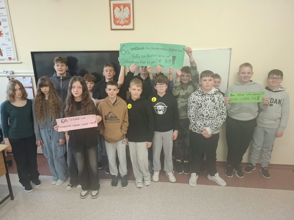 Dzieci stoją wspólnie na tle tablicy szkolnej, trzymając plakaty wykonane na różowych i zielonych kartkach. Hasła podkreślają, jak ważna jest widoczność pieszych na drodze, szczególnie po zmroku.
Na jednym z plakatów można przeczytać:
„6A dobrze wie – odblask chroni Ciebie i mnie!”
Uczniowie wykazali się kreatywnością i zaangażowaniem, tworząc rymowane hasła, które w prosty i przystępny sposób przypominają o zasadach bezpieczeństwa.