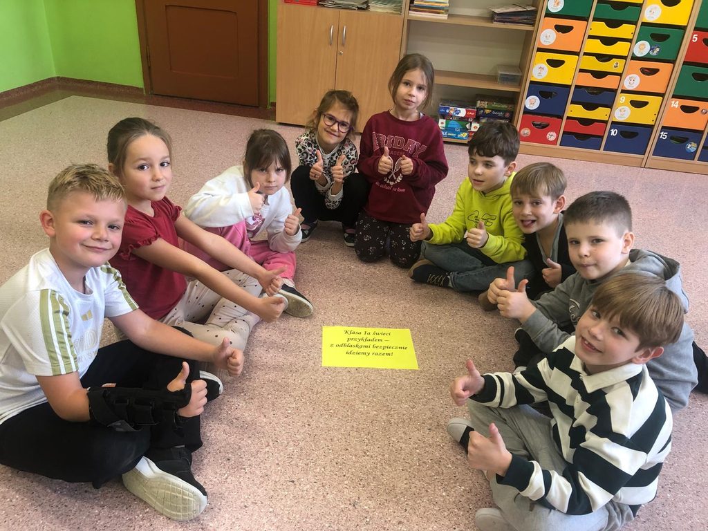 Na zdjęciu widoczni są uczniowie klasy I A, którzy siedzą w kole na podłodze sali lekcyjnej. Dzieci z uśmiechami prezentują gest uniesionego kciuka, wyrażając radość i zadowolenie ze wspólnego działania.

W centrum grupy znajduje się jasnożółta kartka z hasłem:

„Klasa 1a świeci przykładem – z odblaskami bezpiecznie idziemy razem!”
Zdjęcie przedstawia atmosferę współpracy, integracji i wspólnego zaangażowania w promowanie bezpieczeństwa na drodze.