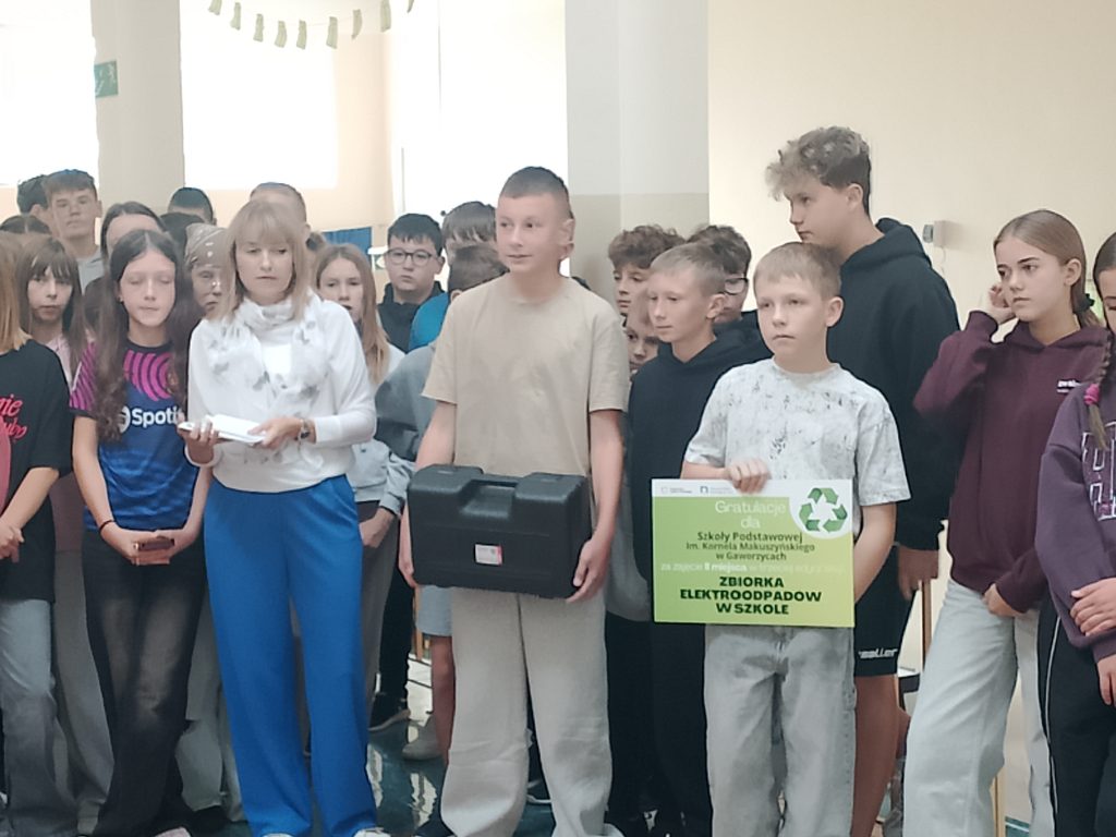 Na zdjęciu uwieczniono uroczyste podsumowanie szkolnej akcji zbiórki elektroodpadów, podczas którego społeczność szkolna świętowała wspólny sukces.
Na pierwszym planie widoczna jest nauczycielka w białej bluzce i niebieskich spodniach, stojąca obok uczniów. Jeden z chłopców trzyma czarną walizkę ze sprzętem, a drugi prezentuje pamiątkową tablicę informującą o zajęciu III miejsca w zbiórce elektroodpadów przez Szkołę Podstawową im. Kornela Makuszyńskiego w Gaworzycach. W tle zgromadzili się pozostali uczniowie, uczestniczący w szkolnym wydarzeniu.