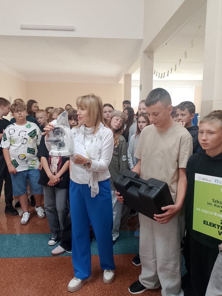Na zdjęciu uwieczniono uroczyste wydarzenie szkolne związane z podsumowaniem akcji zbiórki elektroodpadów.
W szkolnym holu zgromadzili się uczniowie wraz z nauczycielką. Na pierwszym planie widać kobietę prezentującą jeden z przedmiotów oraz ucznia trzymającego czarną walizkę ze sprzętem. Pozostali uczniowie z zainteresowaniem obserwują wydarzenie.
Po prawej stronie widoczna jest zielona tablica informująca o sukcesie szkoły w zbiórce elektroodpadów.