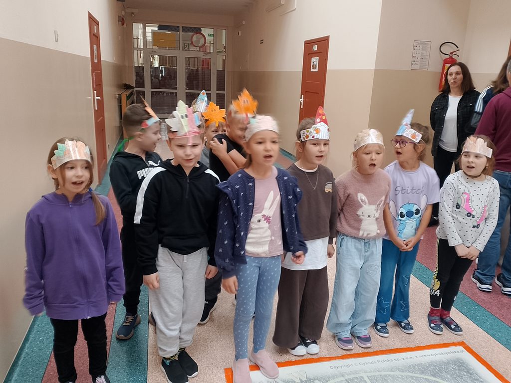 Na zdjęciu widzimy grupę uczniów prezentujących własnoręcznie wykonane, kolorowe nakrycia głowy. Dzieci stoją razem na szkolnym korytarzu, z zaangażowaniem uczestnicząc we wspólnej prezentacji. Różnorodne korony i opaski ozdobione jesiennymi motywami podkreślają kreatywność i pomysłowość uczestników.