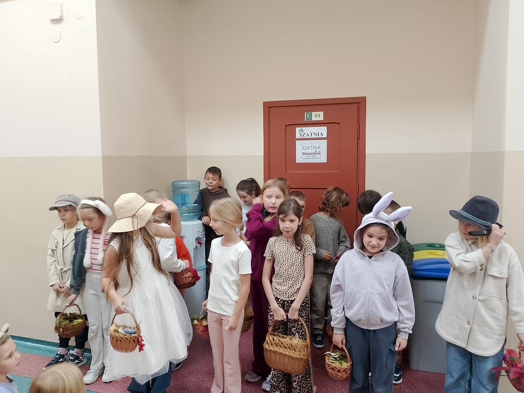Na zdjęciu widzimy grupę uczniów przygotowanych do występu lub szkolnej inscenizacji. Dzieci mają na sobie różnorodne stroje i rekwizyty – kapelusze, opaski z uszami oraz koszyczki – które nadają wydarzeniu wyjątkowy, teatralny charakter.
Uczniowie oczekują na rozpoczęcie prezentacji, a ich stroje wskazują, że każdy z nich wciela się w określoną rolę. Zdjęcie ukazuje zaangażowanie dzieci, kreatywność oraz radosną atmosferę wspólnego szkolnego przedsięwzięcia.