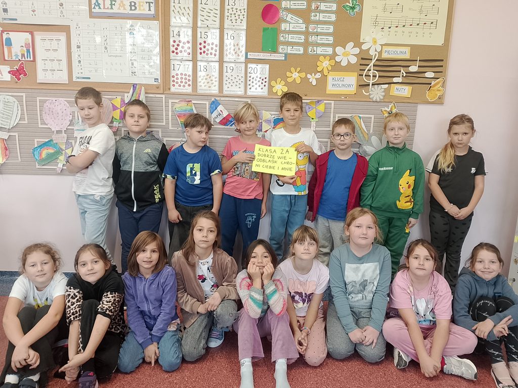 Na zdjęciu widoczni są uczniowie klasy II A, którzy wspólnie pozują do pamiątkowej fotografii w swojej sali lekcyjnej. Dzieci ustawione są w dwóch rzędach – część stoi, a pozostali siedzą na podłodze.

Na środku grupy uczniowie prezentują żółtą kartkę z hasłem:
„Klasa 2A dobrze wie – odblask chroni Ciebie i mnie!”
W tle znajduje się kolorowa dekoracja sali, pełna prac plastycznych, pomocy dydaktycznych oraz elementów związanych z muzyką i nauką. Fotografia przedstawia wspólne zaangażowanie uczniów w promowanie bezpieczeństwa na drodze.