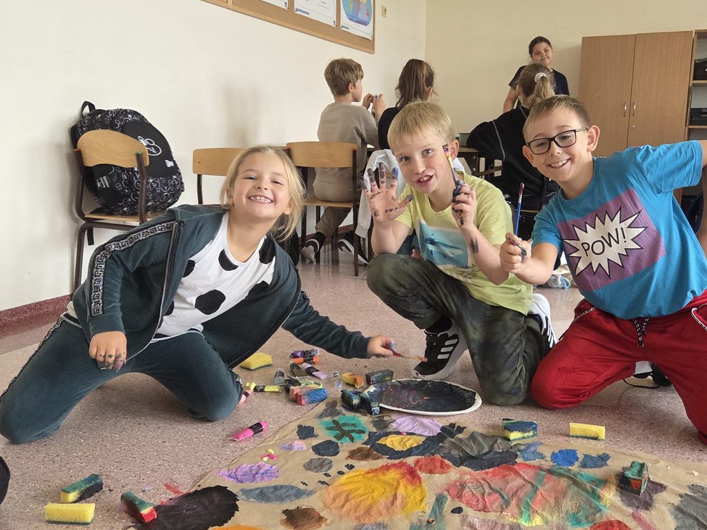 Na pierwszy tle widać dwóch chłopców i dziewczynkę. Wszyscy są uśmiechnięci, pokazują do obiektywu pędzle i dłonie, na których widać ślady farby. Na podłodze rozłożone są tubki z farbami. Dzieci pracują nad malowidłem o tematyce jesiennej. W tle czworo innych uczniów siedzi przy stolikach i jest pogrążonych w rozmowie.