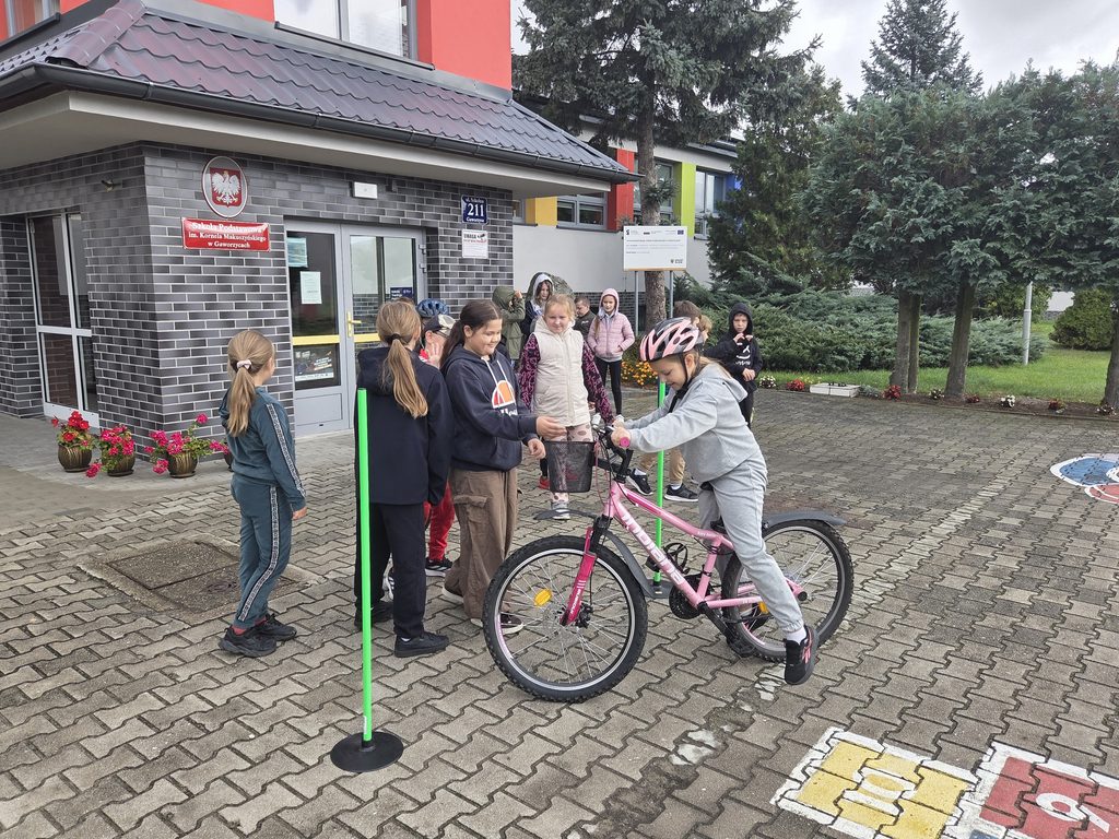 Na pierwszym tle dziewczynka w szarym dresie i różowym kasku siedzi na różowym rowerze. Za nią znajduje się grupka dzieci, czekająca na swoją kolej, by pokonać rowerowy tor przeszkód. Uczniowie stoją przed głównym wejściem do szkoły.