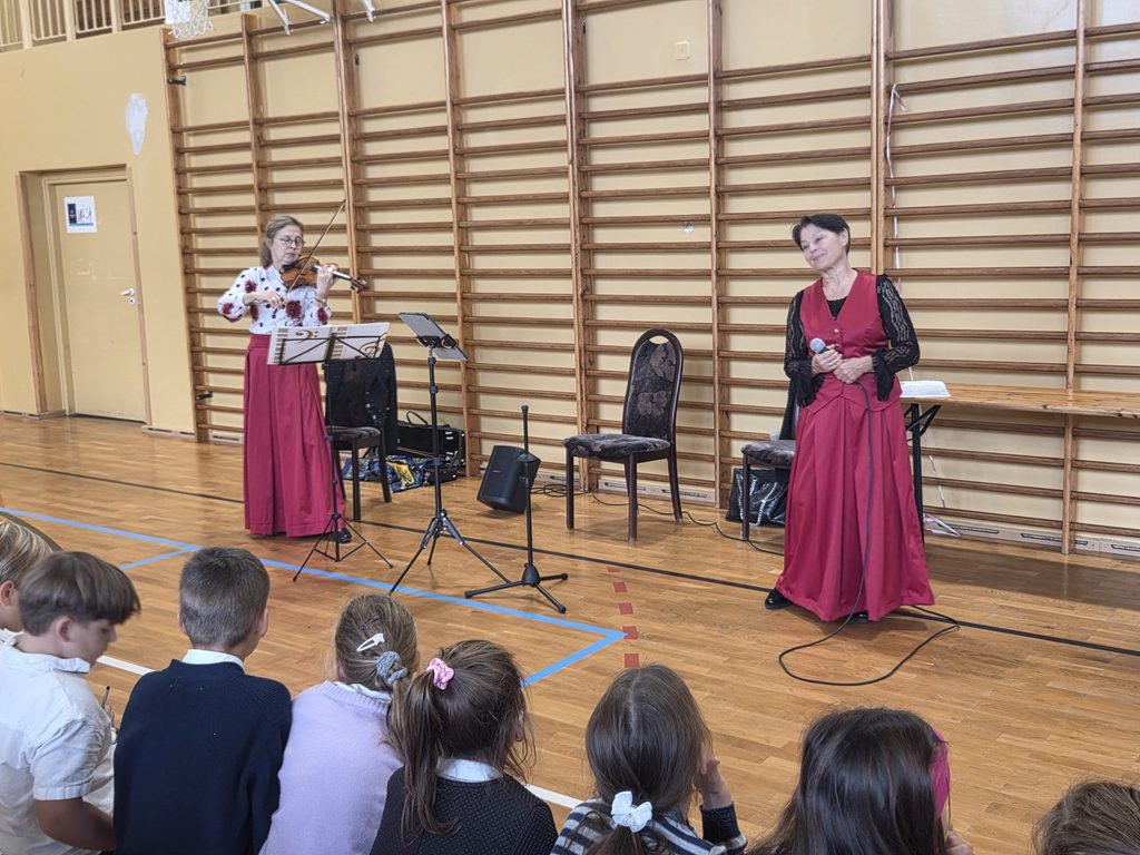 Na zdjęciu widać uczniów oglądających występ muzyczny. Skrzypaczka gra na instrumencie, a prowadząca koncert trzyma mikrofon i przygotowuje się do dalszej części spotkania.
