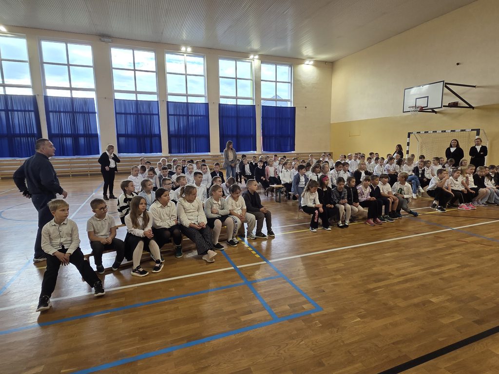 Uczniowie klas 1-3 znajdują się w sali gimnastycznej. Siedzą na drewnianych ławkach - gotowi do uczestniczenia w koncercie. Wyglądają elegancko - mają na sobie białe koszule.