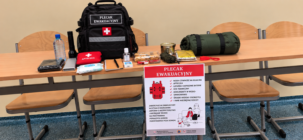 Na zdjęciu przedstawiono pokazowy plecak ewakuacyjny wraz z wyposażeniem, przygotowany w ramach Krajowego Treningu Rodzinnego – Akademii Ochrony Ludności i Obrony Cywilnej (OLiOC).
Na stole zaprezentowano czarny plecak ewakuacyjny oraz podstawowe przedmioty niezbędne w sytuacjach kryzysowych, m.in. wodę pitną, żywność o długim terminie przydatności, apteczkę pierwszej pomocy, latarkę, śpiwór, koc termiczny, środki higieniczne, powerbank i inne przydatne akcesoria. 