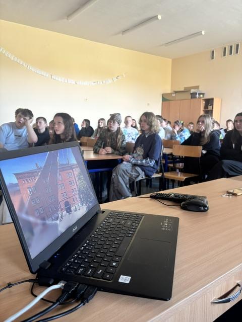 Na zdjęciu widać jasną salę lekcyjną, w której uczniowie siedzą w ławkach i oglądają film edukacyjny wyświetlany na ekranie, podczas spotkania z funkcjonariuszami Policji. Na biurku znajduje się laptop, który służy do odtwarzania materiału. Na końcu sali stoi szafa. Pomieszczenie jest dobrze oświetlone i panuje w nim przyjazna atmosfera sprzyjająca nauce.