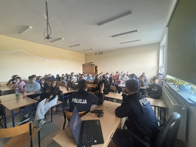Na zdjęciu widać jasną salę, w której odbywa się spotkanie uczniów z policjantami. Sala jest jasna, z dużymi oknami, zielonymi roletami oraz szafami stojącymi przy tylnej ścianie. Uczniowie siedzą na krzesłach w ławkach, zwróceni w stronę funkcjonariuszy, i uważnie ich słuchają. Policjanci siedzą przy biurku, tyłem do osoby robiącej zdjęcie, ale przodem do uczniów. Spotkanie ma charakter edukacyjny i służy przekazaniu młodzieży ważnych informacji dotyczących bezpieczeństwa.